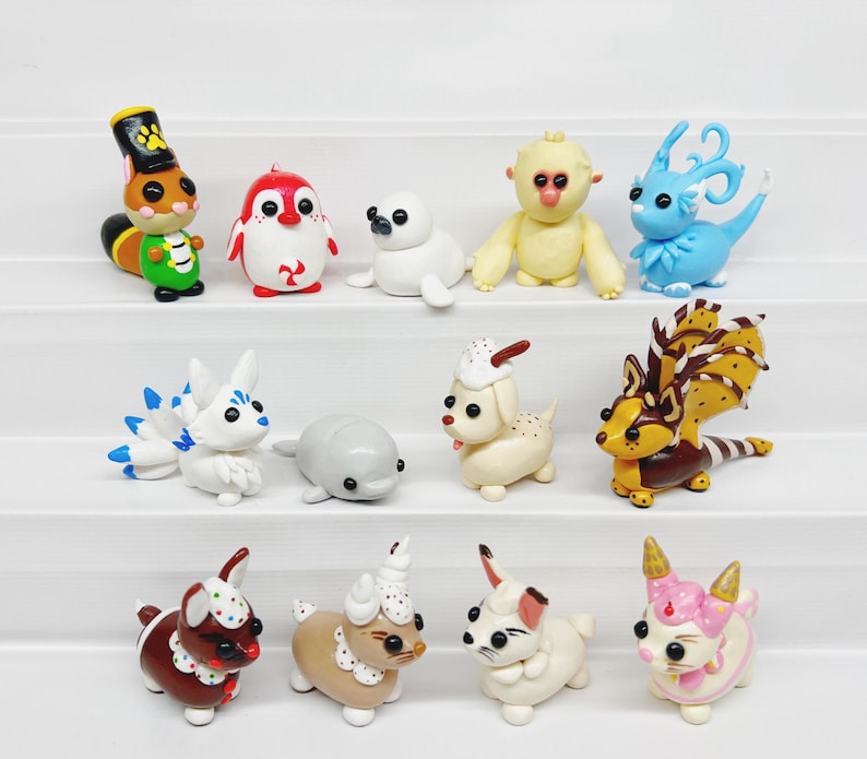 May include: A collection of 14 handmade polymer clay figurines, each depicting a different animal. The figurines are painted in various colors, including white, red, blue, brown, and yellow. The animals include a squirrel, penguin, seal, monkey, dragon, fox, dolphin, dog, deer, and rabbit.