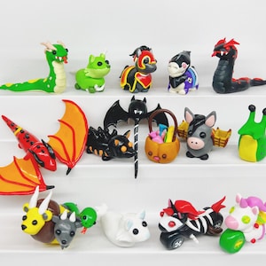 May include: A collection of 14 handmade polymer clay figurines, including a dragon, a bat, a snail, a unicorn, a donkey, a dragon, a bat, a snail, a unicorn, a donkey, a dragon, a bat, a snail, a unicorn, and a donkey. The figurines are painted in various colors, including red, green, black, white, and purple.