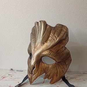 Könnte beinhalten: Eine handgefertigte, goldfarbene Maske mit einem strukturierten, organischen Design. Die Maske hat eine spitze, schnabelartige Nase und Augenöffnungen, mit einem schwarzen Band zum Tragen. Die Oberfläche der Maske hat ein verwittertes Aussehen.