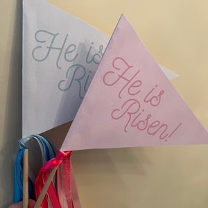 Könnte beinhalten: Zwei dreieckige Papierflaggen mit Holzstöcken und bunten Bändern. Die Flaggen sind rosa und weiß und haben den Text "He is Risen!" in Kursivschrift.