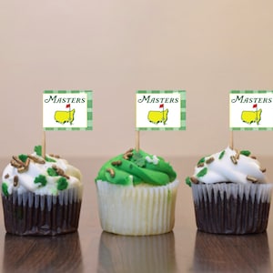 May include: Three cupcakes decorated with green and white frosting and gold sprinkles. Each cupcake has a toothpick with a green and white flag that says "Masters" with a yellow and red golf flag on it.
