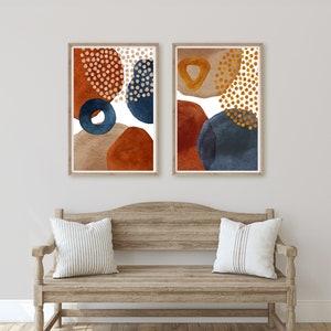 May include: Two framed abstract art prints. Each print features watercolor shapes in rust, navy, tan, and gold. The art is displayed above a wooden bench with striped pillows. The artwork adds a modern touch to the room.