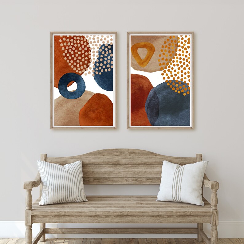 May include: Two framed abstract art prints. Each print features watercolor shapes in rust, navy, tan, and gold. The art is displayed above a wooden bench with striped pillows. The artwork adds a modern touch to the room.