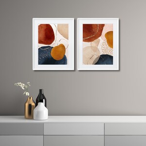 May include: Two framed abstract art prints. Each print features organic shapes in shades of rust, navy, gold, and beige, with gold glitter accents. The prints are displayed in white frames, creating a modern and artistic aesthetic.