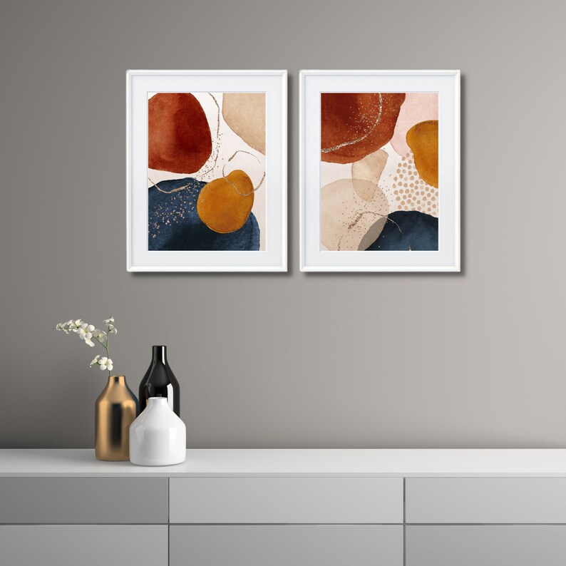 May include: Two framed abstract art prints. Each print features organic shapes in shades of rust, navy, gold, and beige, with gold glitter accents. The prints are displayed in white frames, creating a modern and artistic aesthetic.