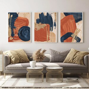 May include: Three abstract art prints with a modern minimalist design. The prints feature bold brushstrokes in shades of burnt orange, navy blue, and gold on a cream background.