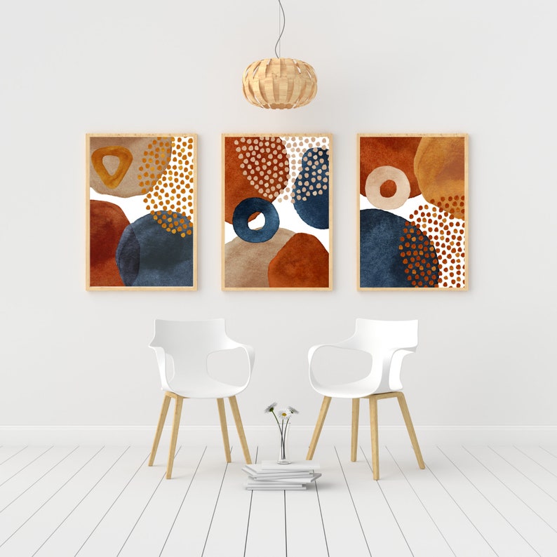 May include: Three framed abstract art prints with a minimalist design. Each print features watercolor-style shapes in rust, navy, tan, and gold. The artwork is displayed above two white chairs with wooden legs, creating a modern interior design.