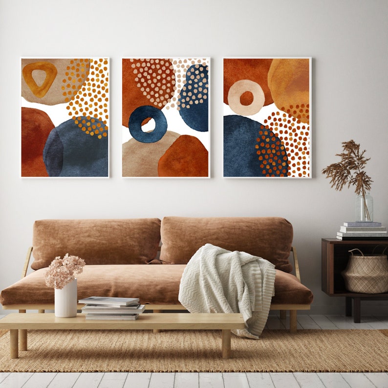 May include: Three framed abstract art prints. Each print features watercolor shapes in shades of rust, navy blue, and beige, with gold and white accents. The artwork is displayed above a brown sofa in a modern living room setting.