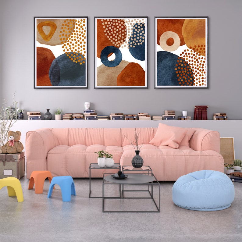 May include: Three framed abstract art prints. Each print features watercolor-style shapes in rust, navy, tan, and gold. The prints are displayed above a pink sofa in a modern living room setting. The room also includes small colorful stools and a beanbag.