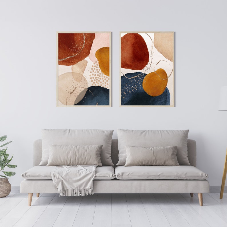 May include: Two framed abstract art prints. Each print features organic shapes in shades of rust, navy, gold, and beige, with gold glitter accents. The artwork is displayed above a light gray sofa with pillows and a blanket.