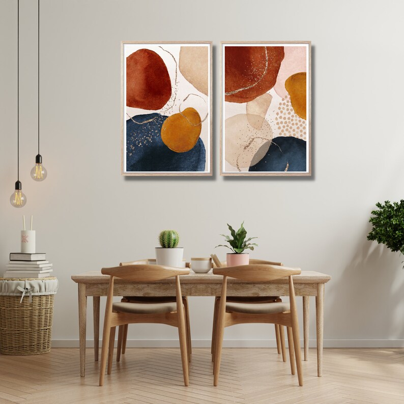 May include: Two framed abstract art prints. Each print features watercolor-style shapes in rust, navy, gold, and beige, with gold metallic accents. The prints are displayed above a wooden dining table with chairs.
