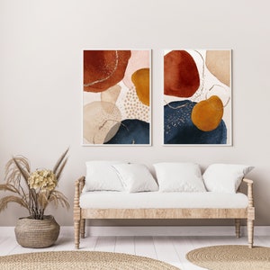 May include: Two framed abstract art prints. Each print features watercolor-style shapes in rust, navy, gold, and beige, with gold glitter accents. The artwork is displayed above a light wood sofa with white cushions.
