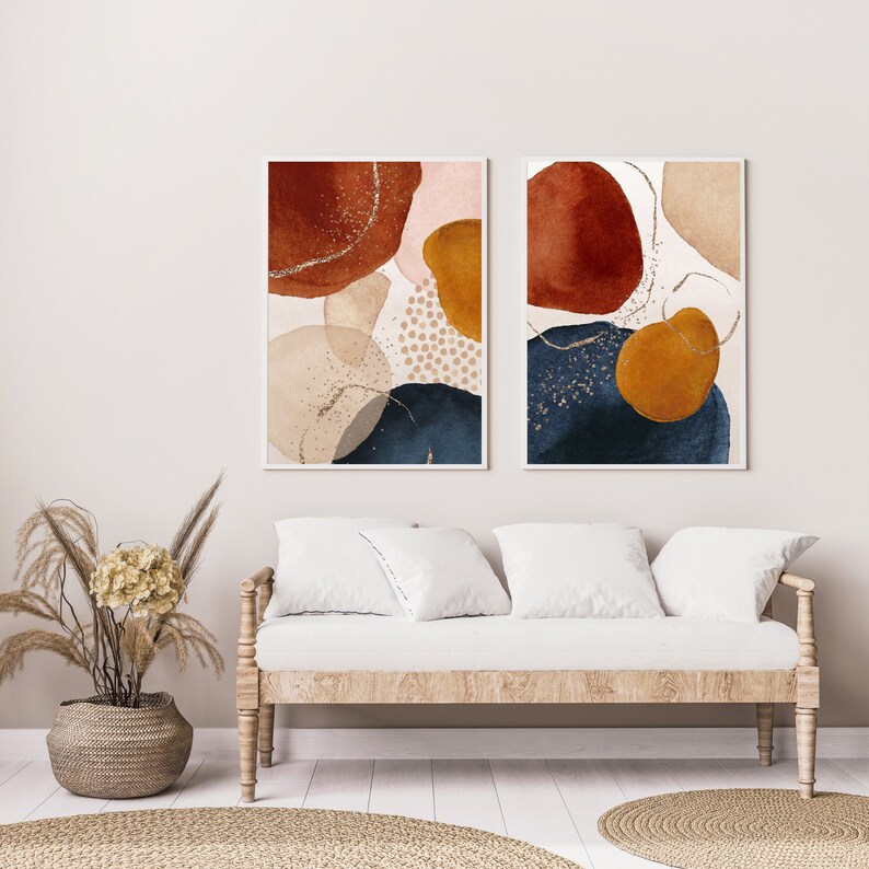 May include: Two framed abstract art prints. Each print features watercolor-style shapes in rust, navy, gold, and beige, with gold glitter accents. The artwork is displayed above a light wood sofa with white cushions.