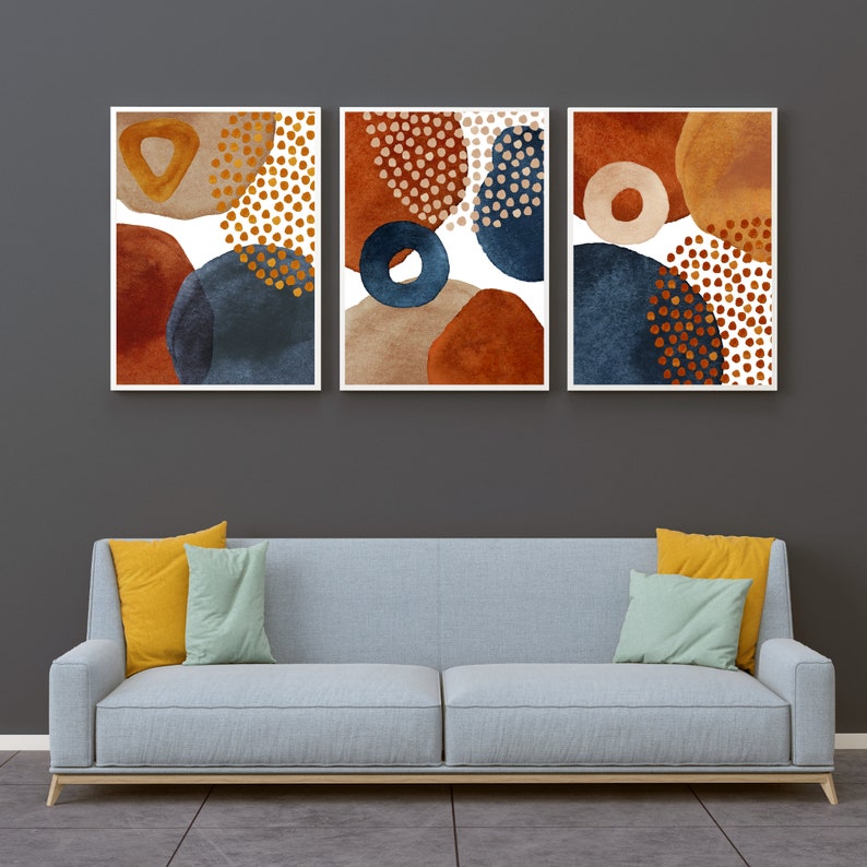 May include: A triptych of abstract art prints featuring watercolor-style shapes in shades of rust, navy blue, and tan, with gold dot accents. The artwork is framed in white and hangs above a light blue sofa with yellow and green pillows.