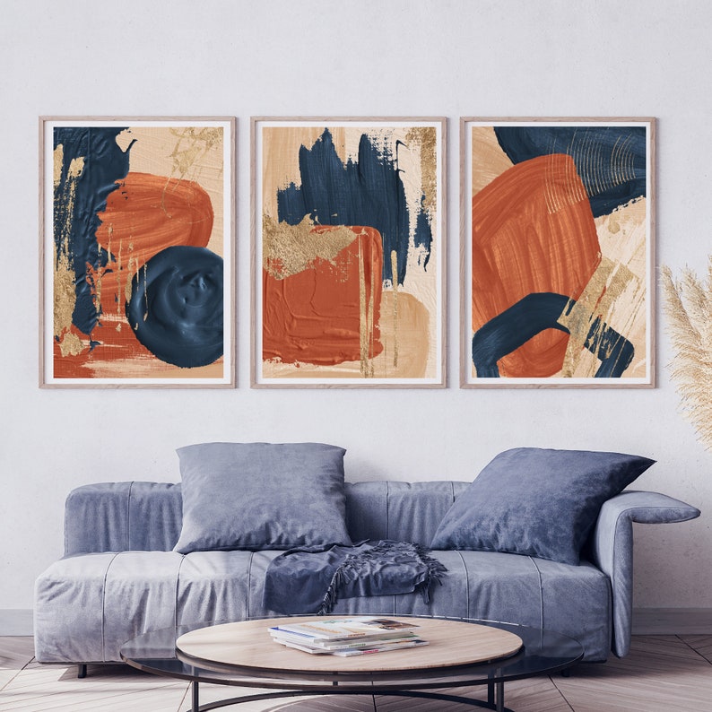 May include: Three abstract art prints with a modern minimalist design. The prints feature bold strokes of blue, orange, and gold paint on a cream background.