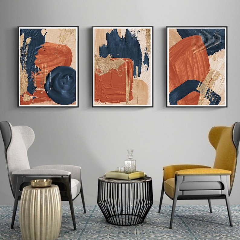 May include: Three abstract paintings with a modern minimalist design. The paintings feature bold strokes of blue, orange, and gold paint on a beige background. The paintings are framed in black and are hung on a gray wall.