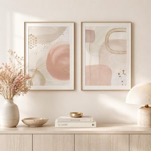 May include: Two framed abstract art prints. Each print features soft pink, beige, and gold watercolour shapes. The artwork is displayed on a light-coloured wall above a wooden cabinet. A vase of dried flowers and decorative items are in the foreground.