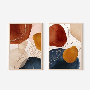 May include: Two framed abstract art prints. Each print features watercolor shapes in rust, navy, mustard, and blush tones, with gold glitter accents. The art has a neutral background and is framed in light wood.