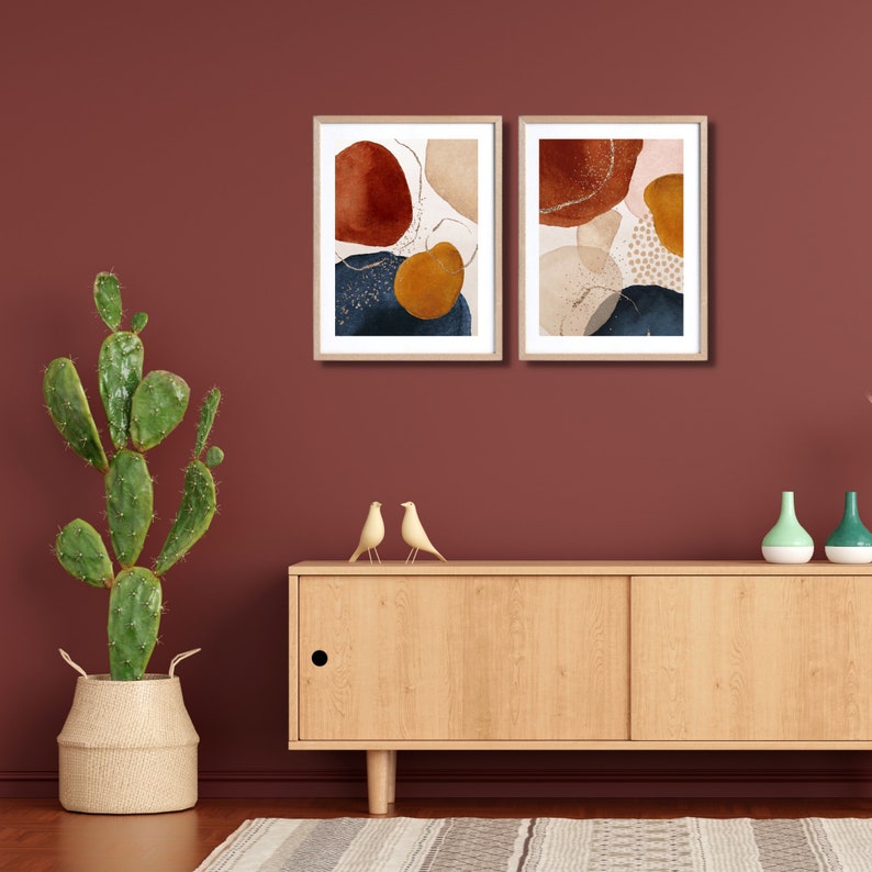May include: Two framed abstract art prints featuring organic shapes in rust, navy, gold, and beige tones. The artwork is displayed on a deep red wall above a wooden cabinet with decorative birds and vases. A cactus in a woven basket is on the left.