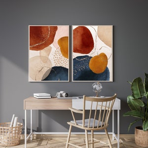 May include: Two framed abstract art prints. Each print features watercolor-style shapes in rust, navy, gold, and beige tones, with gold glitter accents. The artwork is displayed above a wooden desk with a chair and a potted plant.