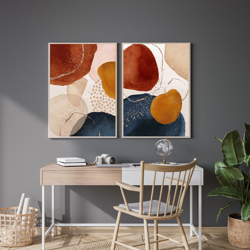 May include: Two framed abstract art prints. Each print features watercolor-style shapes in rust, navy, gold, and beige tones, with gold glitter accents. The artwork is displayed above a wooden desk with a chair and a potted plant.
