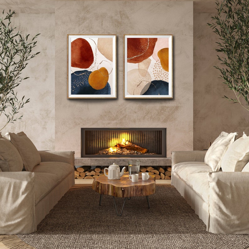 May include: Two framed abstract art prints. Each print features organic shapes in shades of rust, navy, gold, and beige, with gold accents. The artwork is displayed above a fireplace, creating a warm and inviting atmosphere. The room includes neutral-toned sofas and a wooden coffee table.
