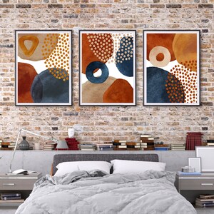 May include: Three framed abstract art prints. Each print features a unique arrangement of geometric shapes in shades of rust, navy, beige, and gold, with dotted and solid patterns. The artwork is displayed against a brick wall.
