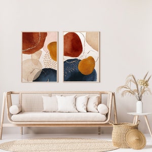 May include: Two framed abstract art prints. Each print features organic shapes in rust, navy, gold, and beige tones with gold accents. The artwork hangs above a light wood sofa with white pillows, a round rug, and a woven basket.