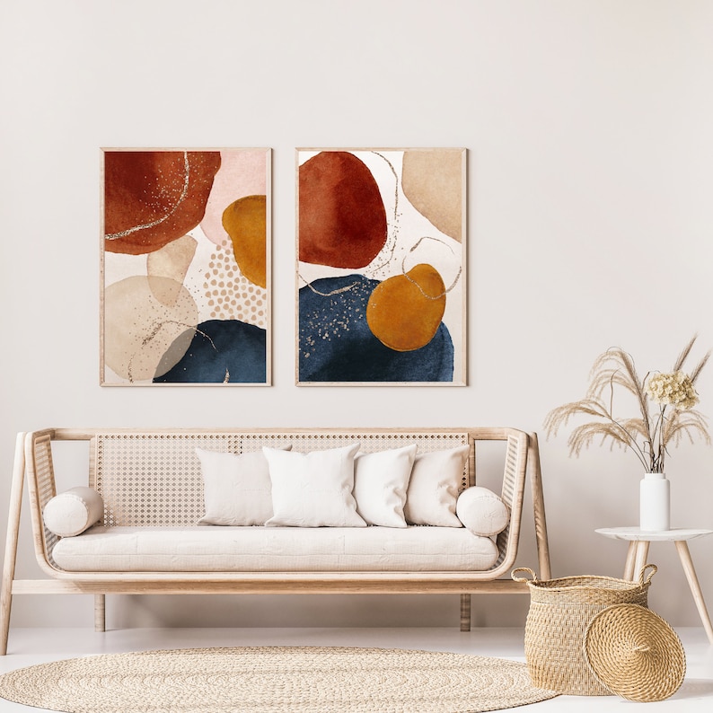 May include: Two framed abstract art prints. Each print features organic shapes in rust, navy, gold, and beige tones with gold accents. The artwork hangs above a light wood sofa with white pillows, a round rug, and a woven basket.