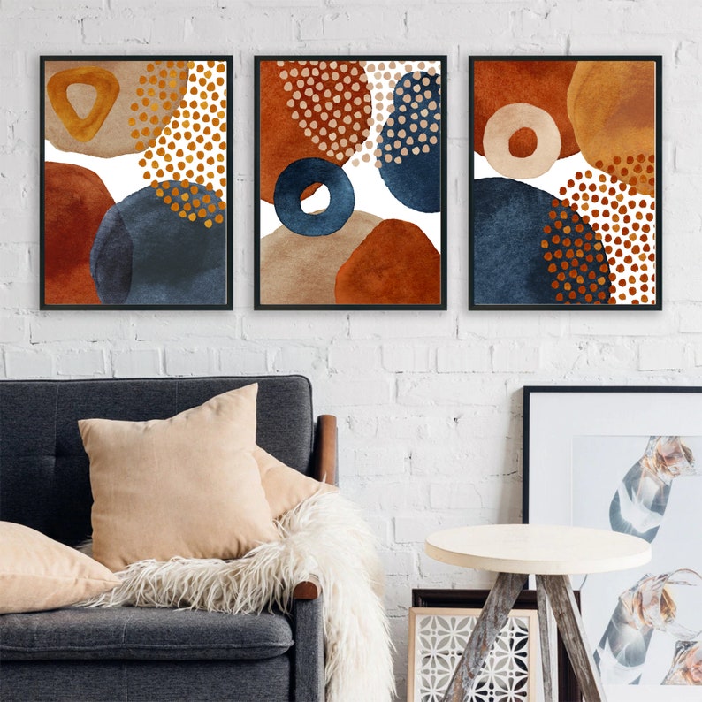 May include: Three framed abstract art prints. Each print features a unique arrangement of watercolor shapes in shades of rust, navy, and beige, with gold dot accents. The prints are displayed on a white brick wall above a gray sofa.