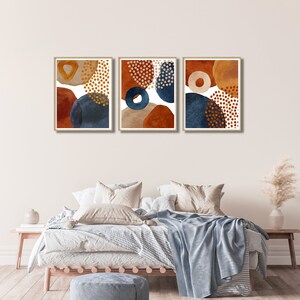 May include: Three framed abstract art prints. Each print features a unique arrangement of organic shapes in shades of rust, navy blue, and beige, accented with gold dot patterns. The artwork is displayed above a bed with neutral bedding.