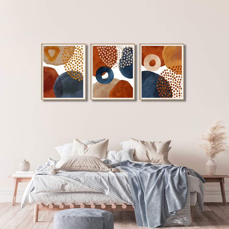 May include: Three framed abstract art prints. Each print features a unique arrangement of organic shapes in shades of rust, navy blue, and beige, accented with gold dot patterns. The artwork is displayed above a bed with neutral bedding.