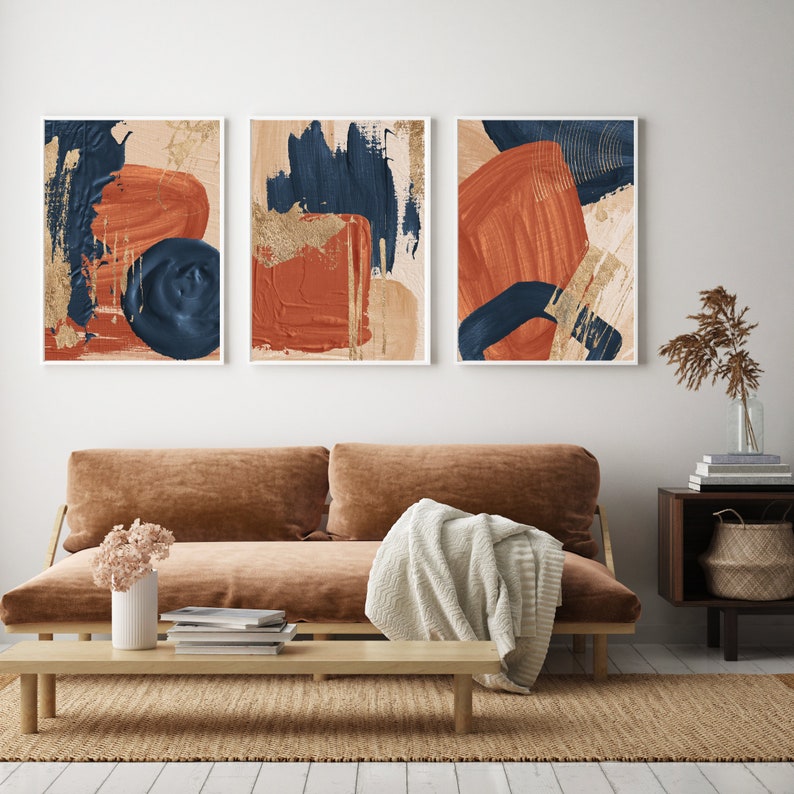 May include: Three abstract art prints with a modern minimalist design. The prints feature bold strokes of blue, orange, and gold paint on a beige background.