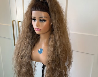 May include: A long, wavy, light brown wig with a high ponytail. The wig is styled with a side part and bangs.
