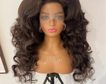 May include: A long, dark brown curly wig on a mannequin head. The wig is styled with a high bouffant and loose curls. The mannequin is on a black tripod stand. The text "JD Wigs" is visible on the mannequin's chest.