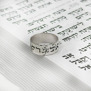 May include: A silver ring with Hebrew text, resting on a book with Hebrew script. The ring has a hammered texture and the text is cut out. The background is a stack of white pages with green text.