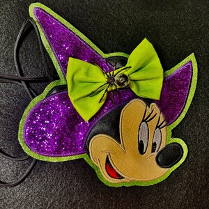 May include: A Halloween-themed Minnie Mouse-shaped crossbody bag. The bag features a purple glitter witch hat with a green bow and a spider embellishment. The face is tan with black and red accents, and a black strap.