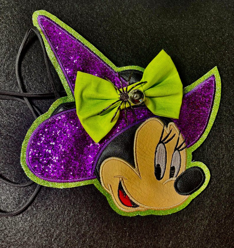 May include: A Halloween-themed Minnie Mouse-shaped crossbody bag. The bag features a purple glitter witch hat with a green bow and a spider embellishment. The face is tan with black and red accents, and a black strap.