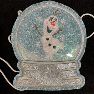 Puede incluir: Un bolso con forma de globo de nieve azul claro y brillante con Olaf de Frozen. El bolso tiene una correa de cordón blanco y una base de purpurina plateada. El aplique de Olaf tiene una nariz de zanahoria y brazos de palo. El globo de nieve está lleno de purpurina.