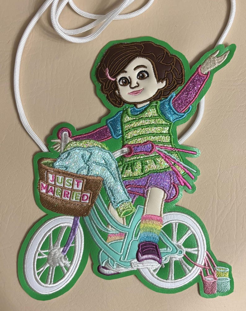 以下が含まれることがあります： 茶色の髪の漫画のキャラクターが水色の自転車に乗っている刺繍ワッペン。自転車には「JUST MARRIED」と書かれた茶色のバスケットが付いています。キャラクターは緑のベスト、紫のショートパンツ、虹色の靴下を着用しており、白いコードが付いています。