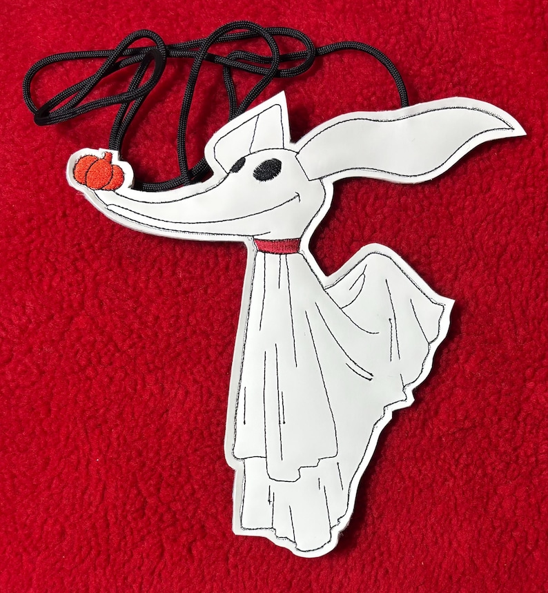 Puede incluir: Un personaje blanco de Zero de The Nightmare Before Christmas, con ojos negros, un collar rojo y una nariz de calabaza. El personaje est&aacute; sujeto a un cord&oacute;n negro. El fondo es rojo liso.