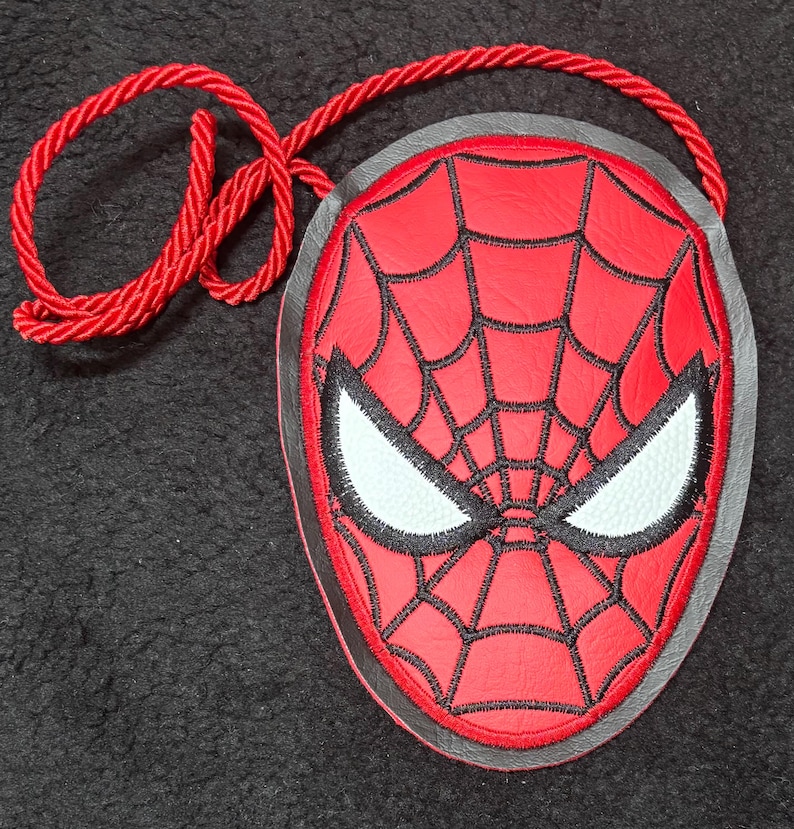 May include: A red and black Spider-Man face-shaped crossbody bag with a red rope strap. The bag features a web design and white eye details. The bag is approximately 7 inches wide.