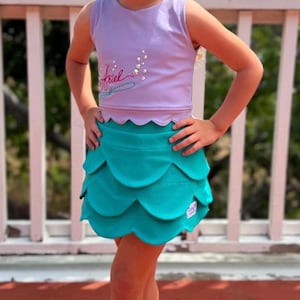 May include: A young girl wearing a lavender tank top with the name "Ariel" and a teal mermaid-scale skirt. The outfit is paired with white sneakers. The girl is smiling and standing on a wooden deck.