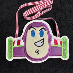 May include: Embroidered Buzz Lightyear face ornament with a red and white striped ribbon. The ornament features a purple helmet, blue eyes, and a smiling expression. The sides have green and red accents.