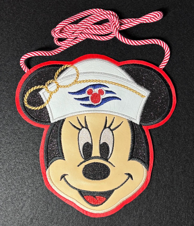 May include: Embroidered Minnie Mouse face with a sailor hat and red trim. The hat features a gold rope design and a Disney Cruise Line logo. A red and white rope is attached for hanging.
