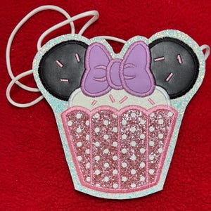 May include: A whimsical cupcake-shaped crossbody bag with black ears and a purple bow. The bag features a pink and white polka-dotted design with glitter accents and a white cord strap. The design is set against a red background.