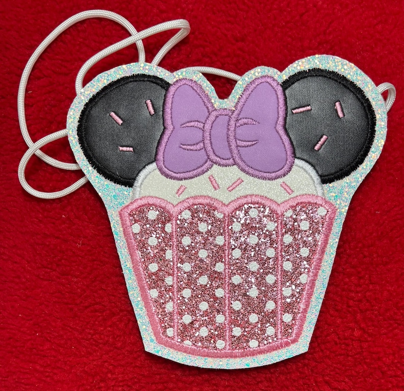 May include: A whimsical cupcake-shaped crossbody bag with black ears and a purple bow. The bag features a pink and white polka-dotted design with glitter accents and a white cord strap. The design is set against a red background.