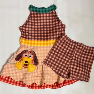 May include: A two-piece children's outfit featuring a sleeveless dress and matching shorts. The dress has a brown and peach plaid bodice, a yellow stripe, and a red scalloped hem. The dress features an applique of a cartoon dog. The shorts are brown and peach plaid.