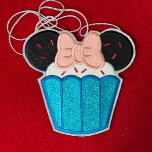 May include: A cupcake-shaped applique with black glitter ears and a pink bow. The cupcake has a blue glitter base and white frosting with pink sprinkles. A silver cord is attached for hanging.