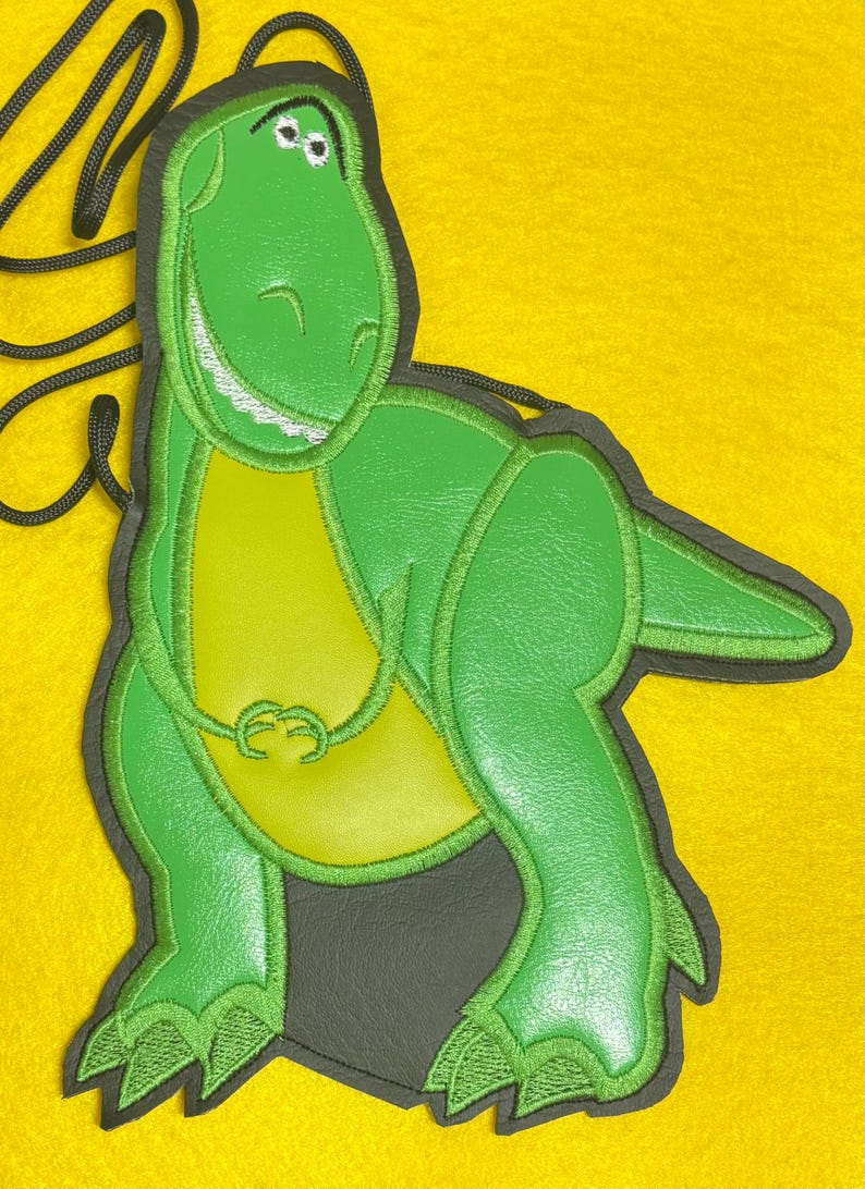 May include: Green dinosaur-shaped bag with a black cord. The bag features a smiling dinosaur design with a light green belly and dark green outline. The background is yellow.